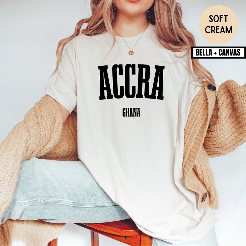 Op de afbeelding: Een zacht cr&egrave;mekleurig T-shirt met het woord "ACCRA" in vette zwarte letters, met "GHANA" eronder gedrukt. Het shirt is van het merk Bella + Canvas. Het model draagt een beige vest en een blauwe jeans.