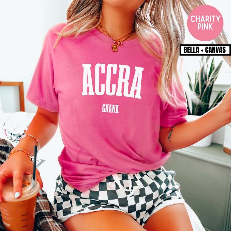 Op de afbeelding: Een felroze t-shirt met het woord "ACCRA" in witte blokletters boven "GHANA". Het shirt is gemaakt van Bella + Canvas materiaal. Het shirt heeft een levendige roze kleur en de tekst is vetgedrukt en gemakkelijk te lezen.