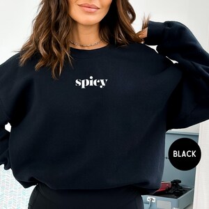 Puede incluir: Sudadera negra con la palabra "spicy" en blanco, centrada en el pecho. La sudadera es de color negro liso. Un c&iacute;rculo negro con la palabra "BLACK" en blanco est&aacute; en la esquina inferior derecha.