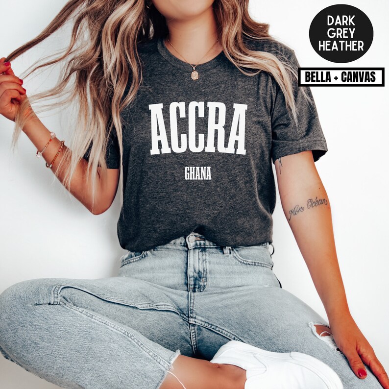 Op de afbeelding: Donkergrijze heather t-shirt met het woord "ACCRA" in dikke witte letters boven "GHANA". Het shirt is van het merk Bella + Canvas.