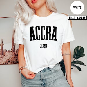 Op de afbeelding: Wit T-shirt met het woord "ACCRA" in dikke zwarte letters boven "GHANA". Het shirt is van het merk Bella + Canvas. Het model draagt een blauwe spijkerbroek en armbanden.