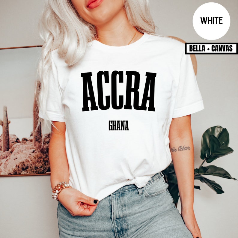 Op de afbeelding: Wit T-shirt met het woord "ACCRA" in dikke zwarte letters boven "GHANA". Het shirt is van het merk Bella + Canvas. Het model draagt een blauwe spijkerbroek en armbanden.