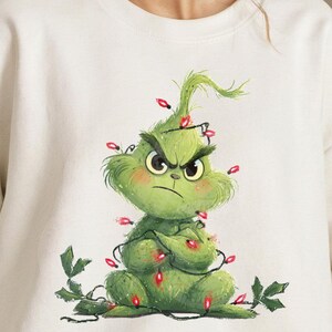 Peut inclure: Sweat-shirt blanc avec un motif du Grinch vert. Le Grinch affiche une expression fâchée, entouré de guirlandes lumineuses rouges. Le motif comprend des feuilles vertes, sur le thème des fêtes de fin d'année.