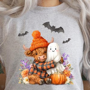 May include: A light gray sweatshirt featuring a Halloween-themed graphic. The design includes a Highland cow wearing an orange knit hat and plaid scarf, hugging a ghost. A small pumpkin and bats are also included in the design.
