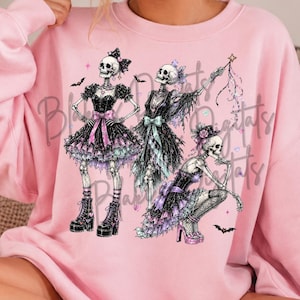 May include: A light pink crewneck sweatshirt featuring a graphic print of three skeletons wearing black and purple ruffled dresses, bows, and platform boots, with small bats and stars surrounding the figures in a whimsical, dark-themed illustration.