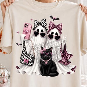 May include: A cream-colored t-shirt featuring a graphic of two white ghosts and a black cat. The ghosts wear heart-shaped sunglasses and bows, one with a zebra print and one with a pink leopard print. The cat wears pink glasses and a pearl collar.
