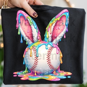 May include: Black sweatshirt featuring a baseball with bunny ears. The baseball is white with red stitching, and the bunny ears are pink and white. The design has colorful, dripping paint in shades of blue, yellow, green, and pink.