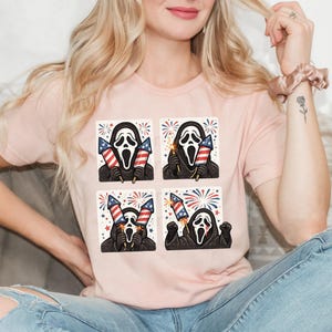 K&ouml;nnte beinhalten: Hellrosa T-Shirt mit einem sich wiederholenden 2x2 Rastermuster. Jedes Feld zeigt die Ghostface-Maske aus den Scream-Filmen, die Feuerwerk in den Farben der amerikanischen Flagge h&auml;lt.