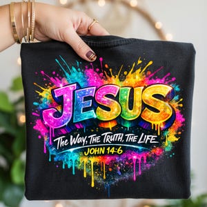 May include: A black t-shirt featuring a vibrant, multi-colored paint splatter design. The word JESUS is printed in large, glittery, rainbow-colored letters. Below, the text reads The Way, The Truth, The Life and the scripture reference JOHN 14:6.