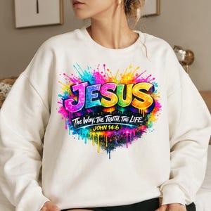 May include: A cream-colored crewneck sweatshirt featuring a vibrant, multi-colored paint splatter design. The graphic displays the word JESUS in bold, glittery letters above the text The Way, The Truth, The Life and John 14:6 in white.