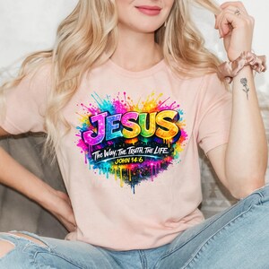 May include: A light peach-colored t-shirt featuring a vibrant, multi-colored paint splatter graphic. The design includes the word JESUS in bold, rainbow-colored letters, followed by the text The Way, The Truth, The Life and JOHN 14:6.