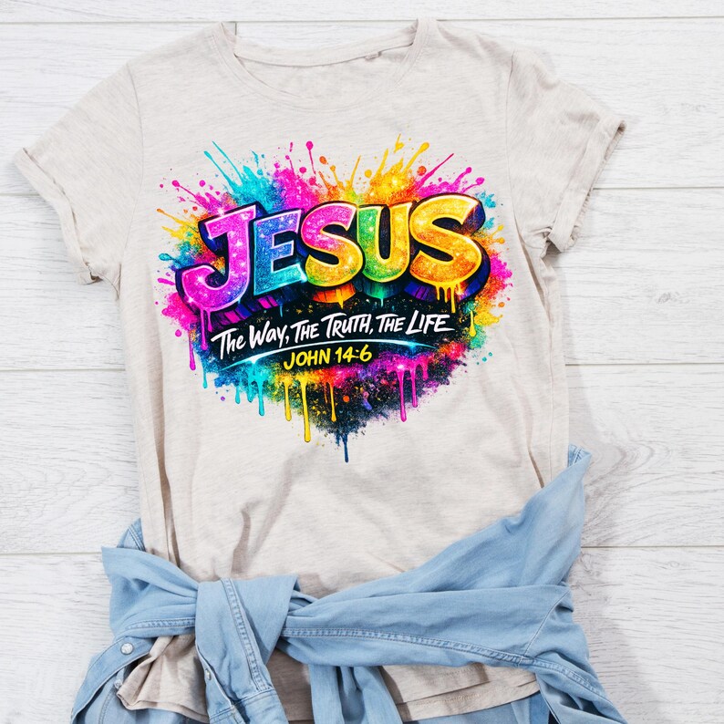 May include: A beige short-sleeve t-shirt featuring a vibrant, multi-colored paint splatter graphic. The design includes the word JESUS in large, bold letters, followed by the text The Way, The Truth, The Life and the reference JOHN 14:6.