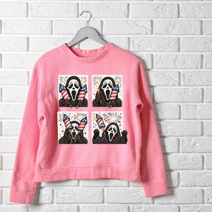 K&ouml;nnte beinhalten: Hellrosa Sweatshirt mit einem sich wiederholenden Muster der Ghostface-Maske aus den Scream-Filmen, die Feuerwerk und amerikanische Flaggen h&auml;lt. Das Design ist in einem Vier-Quadrat-Muster gehalten. Das Sweatshirt h&auml;ngt an einem grauen Kleiderb&uuml;gel.