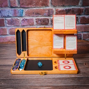 May include: Wooden tabletop gaming box, open to reveal compartments for dice, cards, and accessories. The box is made of light-coloured wood and has a black felt-lined area for rolling dice. Several game cards are visible.
