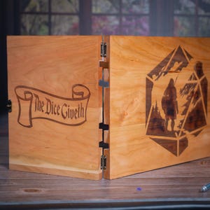 Puede incluir: Una pantalla de maestro de juego de madera con un diseño de rollo en un lado y una imagen estilizada de una persona de pie dentro de un dado grande en el otro lado. El rollo dice "The Dice Giveth".