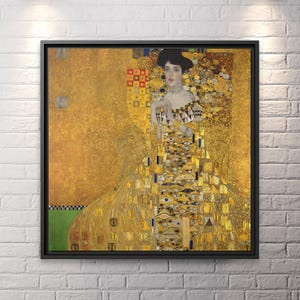 Könnte beinhalten: Gerahmter Druck eines Gemäldes von Gustav Klimt. Das Kunstwerk zeigt eine Frau in einem goldenen Kleid mit komplizierten Mustern. Der Hintergrund ist eine Mischung aus Gold- und Gelbtönen, mit einem schwarzen Rahmen. Das Gemälde wird an einer weißen Backsteinmauer ausgestellt.