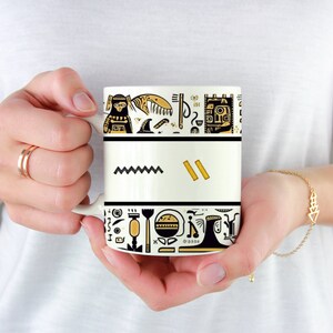 May include: A white ceramic mug with a black and gold design. The design features abstract shapes and symbols, including a zigzag line, rectangles, and other geometric patterns. The mug is held by a person.