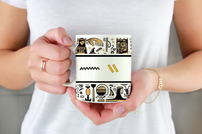 May include: A white ceramic mug with a black and gold design. The design features abstract shapes and symbols, including a zigzag line, rectangles, and other geometric patterns. The mug is held by a person.