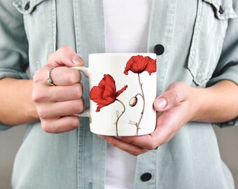 May include: A white ceramic mug featuring two red poppy flower illustrations. The poppies have detailed petals and stems, with a bud visible. The mug is held by a person wearing a light green shirt.