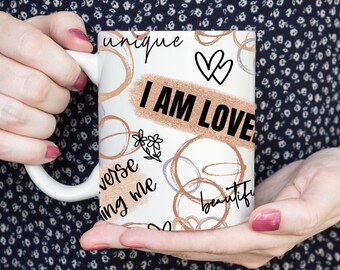 May include: White ceramic mug with the words "I AM LOVE" in black text on a peach-colored background. The mug features a design of abstract circles and words like "unique" and "beautiful".