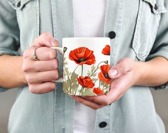 May include: White ceramic mug featuring a vibrant floral design of red poppies with green leaves and stems. The mug has a white handle and is held by a person wearing a light green shirt.