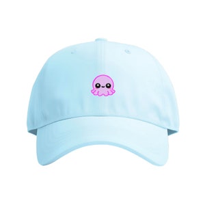 Könnte beinhalten: Eine hellblaue Baseballkappe mit einem gestickten rosa Tintenfisch mit einem lächelnden Gesicht.