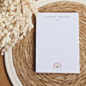 Könnte beinhalten: Ein weißer Notizblock mit dem Text "HAPPY NOTES" und einem kleinen Herzsymbol. Der Notizblock hat ein Regenbogen-Design und den Text "PLOTTING LOVE HERE" unten. Der Notizblock liegt auf einer geflochtenen Strohmatte mit weißem Rand.
