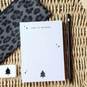 Könnte beinhalten: Ein Schreibwarenset mit einem Notizblock mit dem Text "TIME TO WONDER", einem schwarzen Bleistift, einem kleinen Radiergummi mit Baumgrafik und einem grauen Federmäppchen mit Leopardenmuster. Die Artikel sind auf einem geflochtenen Tablett angeordnet.