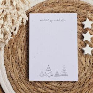 Könnte beinhalten: Ein weißer Notizblock mit dem Text "merry notes" oben. Der Notizblock hat ein gepunktetes Muster und eine Zeichnung von drei Weihnachtsbäumen unten. Der Notizblock liegt auf einer geflochtenen Strohmatte mit drei weißen sternförmigen Dekorationen.
