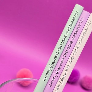 Könnte beinhalten: Drei bunte Strohhalme mit dem Text "ICH BIN (chrerin / WAS SIND DEINE SUPERKRÄFTE?" bedruckt. Die Strohhalme sind in einem Glas auf einem rosa Hintergrund angeordnet.