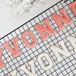 Könnte beinhalten: Nahaufnahme von zwei Reihen gestickter Aufnäher mit der Aufschrift "YVONNE". Die obere Reihe ist korallenfarben und die untere Reihe cremefarben. Die Aufnäher befinden sich auf einem schwarzen Gitter.