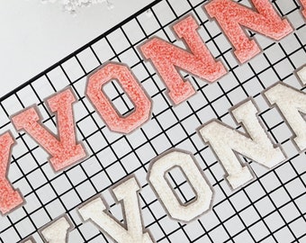 May include: Close-up of two rows of embroidered patches spelling "YVONNE". The top row is in a coral color, and the bottom row is in a cream color. The patches are on a black grid.