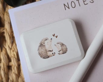 May include: A white, rectangular eraser with a watercolor illustration of two hedgehogs and two small hearts. The eraser sits on a pink notepad with the word "NOTES" printed on it. A white pen is also visible.