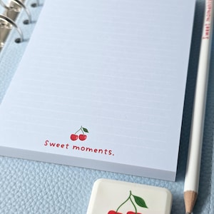 Könnte beinhalten: Hellblauer Notizblock mit Linien, Kirschdesign und dem Text "Sweet moments." Ein weißer Bleistift und ein passender Radiergummi mit Kirschmotiv sind ebenfalls vorhanden. Der Notizblock liegt auf einer hellblauen Oberfläche.