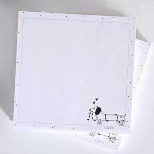 Könnte beinhalten: Quadratischer weißer Notizblock mit gepunktetem Rand und einer schwarz-weißen Dackel-Illustration, die eine Herzkette hält. Das Wort "LOVE" ist darunter gedruckt.