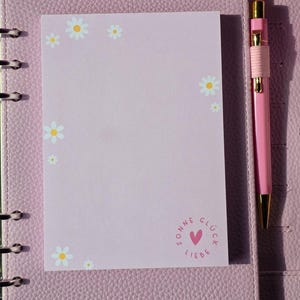 Könnte beinhalten: Ein rosa Notizblock mit Gänseblümchen-Illustrationen und den Worten "Sonne Glück Liebe" in einem Kreis, neben einem rosa Stift mit goldenen Akzenten. Der Notizblock befindet sich in einem rosa lederartigen Ordner.