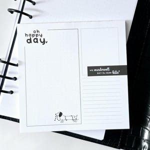 Könnte beinhalten: Ein quadratisches, weißes Notizblatt mit einer schwarzen Strichzeichnung eines Dackels, der mit einer Lichterkette umwickelt ist. Der Text lautet "oh happy day" oben links und "WIE WUNDERVOLL BIST DU DENN bitte" auf einem schwarzen Banner.