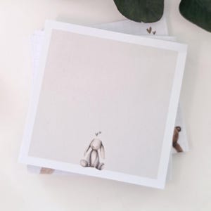 Könnte beinhalten: Ein Stapel quadratischer Notizblöcke mit einer weichen, neutralen Farbpalette. Der oberste Notizblock zeigt eine sitzende Hasenillustration mit Herzen über dem Kopf. Das Design ist schlicht und elegant.
