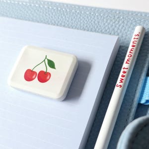 May include: A white eraser with a cherry design and a white pencil with the text "Sweet moments." are on a light blue lined notepad. The notepad is on a light blue leather-like surface.