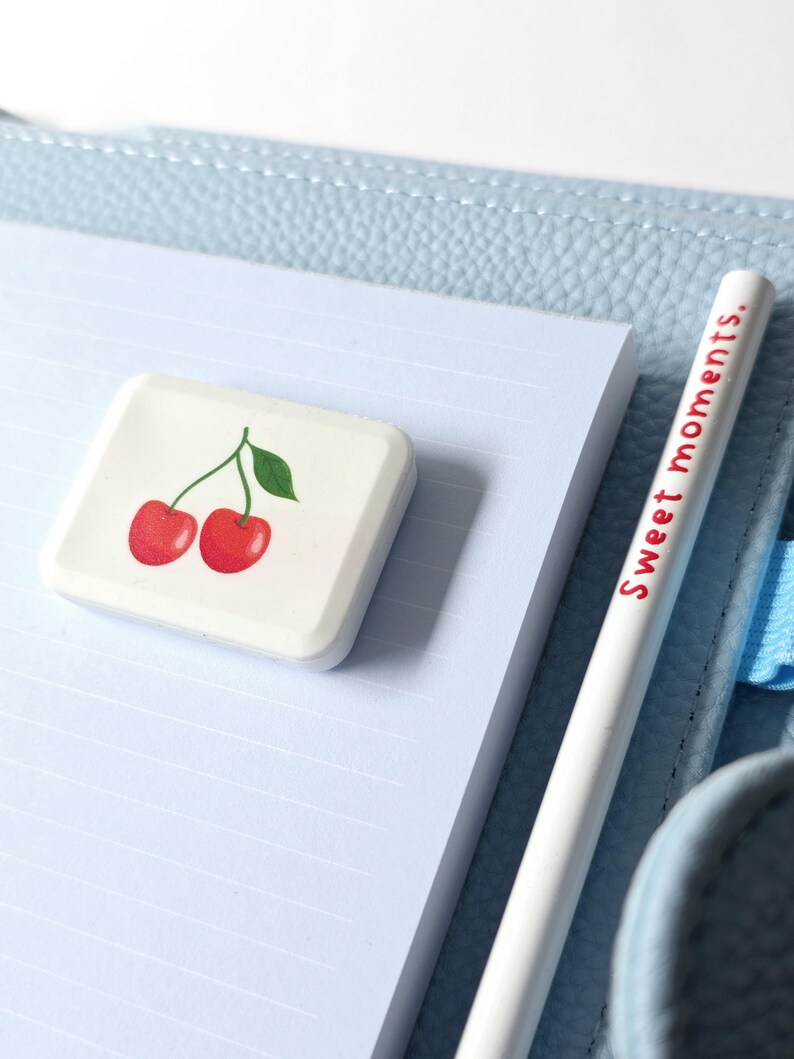 May include: A white eraser with a cherry design and a white pencil with the text "Sweet moments." are on a light blue lined notepad. The notepad is on a light blue leather-like surface.