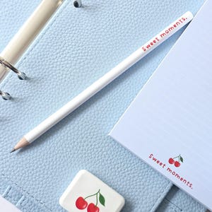 May include: A light blue planner with silver rings, a white pencil, and a notepad. The notepad and eraser feature a cherry design and the words "Sweet moments." The planner is open, revealing lined paper.