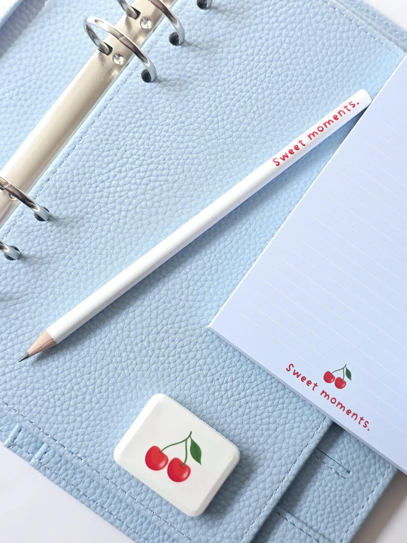 May include: A light blue planner with silver rings, a white pencil, and a notepad. The notepad and eraser feature a cherry design and the words "Sweet moments." The planner is open, revealing lined paper.