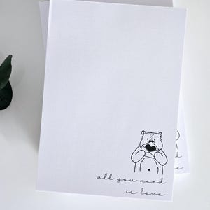 Könnte beinhalten: Weiße Grußkarten mit einer schwarzen Strichzeichnung eines Bären, der ein Herz hält. Unter dem Bären steht in einer Schreibschrift der Satz "all you need is love". Die Karten sind gestapelt.
