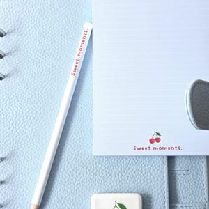 May include: A light blue leather planner with a white pencil and notepad. The pencil and notepad have the text "Sweet moments." A small eraser with a cherry design is also present. The notepad has a cherry graphic.