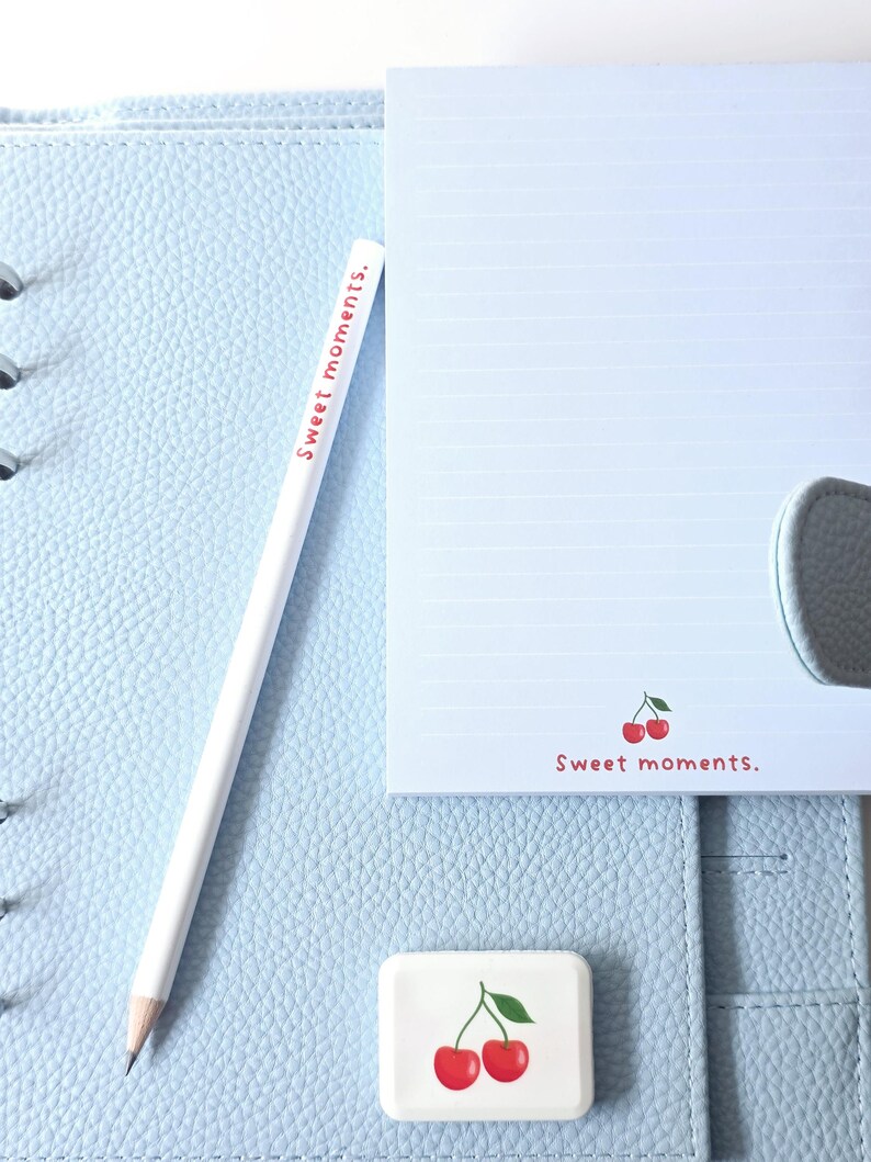 May include: A light blue leather planner with a white pencil and notepad. The pencil and notepad have the text "Sweet moments." A small eraser with a cherry design is also present. The notepad has a cherry graphic.