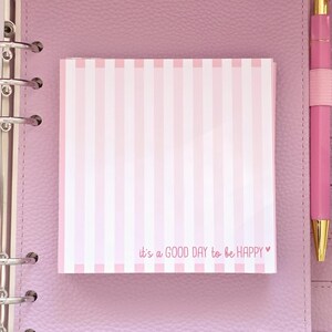 Könnte beinhalten: Ein rosa-weiß gestreifter Notizblock mit dem Text "it's a GOOD DAY to be HAPPY" liegt auf einem rosa Planer. Rechts vom Notizblock befindet sich ein rosa Stift mit goldenen Akzenten. Der Planer hat silberne Ringe.