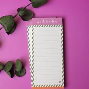 Könnte beinhalten: Rechteckiger Notizblock mit dem Text "my To-Do & Ta da list" in lila Schrift. Der Notizblock hat ein weißes Innenteil mit linierten Seiten und einen Rand aus diagonalen grün-weißen Streifen. Grüner Blätterzweig im Hintergrund.