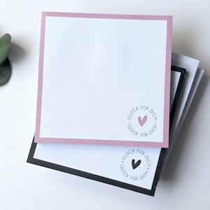 Könnte beinhalten: Stapel quadratischer Notizblöcke mit weißem Papier und farbigen Rändern. Der obere Notizblock hat einen rosa Rand und einen rosa Herzstempel mit den Worten "Glück für dich". Der untere Notizblock hat einen schwarzen Rand und einen schwarzen Herzstempel.