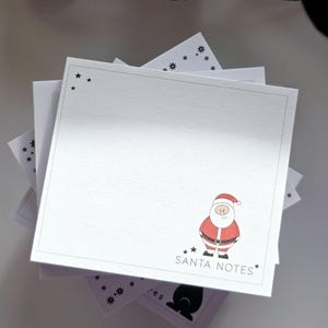 Könnte beinhalten: Ein Stapel weißer quadratischer Notizblöcke mit gepunktetem Rand und Sternakzenten. Jeder Notizblock zeigt eine Cartoon-Illustration des Weihnachtsmanns in Rot, Weiß und Schwarz mit dem Text "SANTA NOTES".