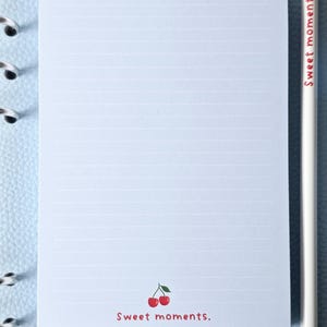 May include: A lined notepad with a cherry illustration and the text "Sweet moments." A white pencil with the text "Sweet moments." is next to the notepad. A small eraser with a cherry illustration is also present.